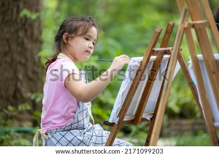 Cute little artist painting picture painting on canvas in nature forest, Happy little girl in garden with brush sitting painting picture , Child artist draws with brush paints on canvas.