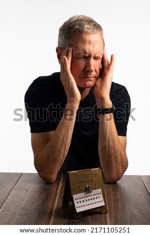 Stressed elderly man eyes shut resting on a dark wood table with a white background