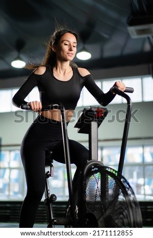 Young Athletic middle aged woman training on air resistance bike, cross training workout set in gym. Active woman spinning a air bike in gym with trainers. female training on air bike. Royalty-Free Stock Photo #2171112855