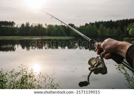 Fisherman with rod, spinning reel on the river bank. Man catching fish, pulling rod while fishing from lake or pond with text space. Fishing for pike, perch on beach lake or pond. Fishing day concept.