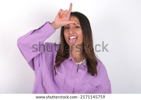 Funny young beautiful caucasian woman wearing pink jacket over white background makes loser gesture mocking at someone sticks out tongue making grimace face.