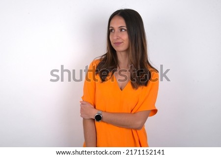 Pleased young beautiful caucasian woman wearing orange T-shirt over white background keeps hands crossed over chest looks happily aside