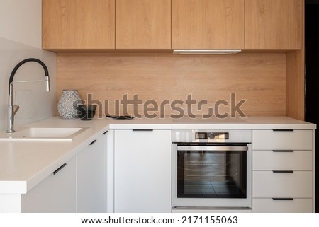Simple kitchen with white cupboards, drawers and countertops, sink, oven and induction hob and wooden backsplash and top cabinets Royalty-Free Stock Photo #2171155063