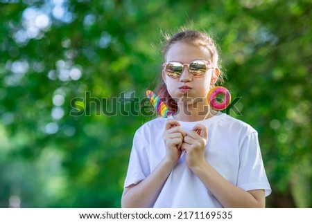 A teenage girl in sunglasses holds lollipops in the form of a pink donut and ice cream. Walking child in the summer park. Horizontal photo of people with space for text.