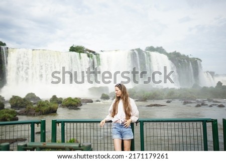 In the photo, a beautiful girl with glasses and white strands in her hair stands against the backdrop of Iguazu Waterfalls located on the border of Brazil (Paraná state) and Argentina 
