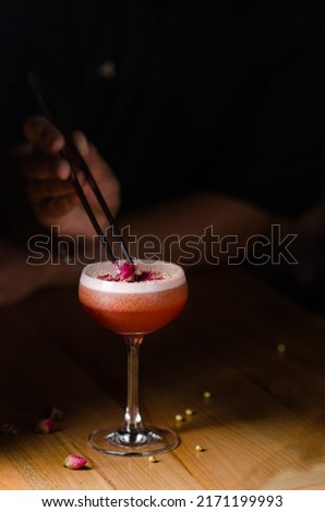 Classic cocktail glass on a wooden table in a nightclub restaurant. Alcoholic cocktail of pink color with a rose, close-up. Modern alcoholic drink. Bartender's hands decorate a cocktail Royalty-Free Stock Photo #2171199993