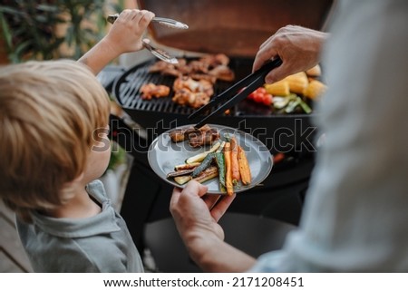 Unrecognizable father with little son grilling ribs and vegetable on grill during family summer garden party, close-up