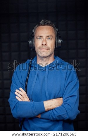 Portrait of male radio host with headphones with arms crossed standing in studio and looking at camera.