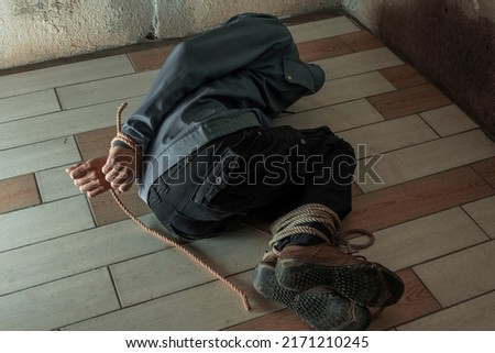 an unrecognizable man in a hood is captured in the basement bound by a rope hand and foot . The kidnapped man is tied up, sitting in the basement. Hands and feet are tied with rope. lying on the floor Royalty-Free Stock Photo #2171210245