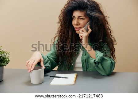 Interested confused tanned curly Latin lady have call from fraudsters sit at the table with notebook isolated over pastel beige background look aside. Copy space Mockup Banner. Concept frauds call