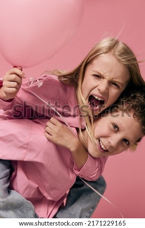 joyful, happy children of school age, on a pink background in pink clothes are playing and the boy is rolling an emotional girl screaming at the camera, with a pink balloon on her back