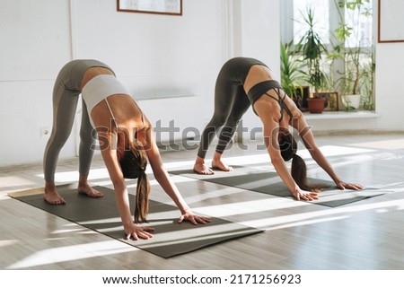 Young fit women practice yoga doing asana in a bright yoga studio. Yoga ticher doing asana sun salutation with student in yoga class Royalty-Free Stock Photo #2171256923