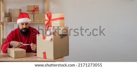 bearded indian man wearing a Santa Claus hat works in a warehouse of boxes with gifts and orders from an online store for Christmas. Small Business Owner Sells Happy New Year. Delivery holiday. banner