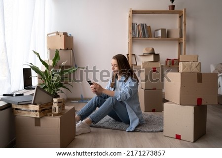 Attractive young woman is moving, sitting among cardboard boxes, using a smartphone and smiling, communicate via smartphone after moving to new flat. Royalty-Free Stock Photo #2171273857