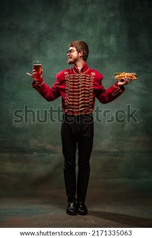 Lunch time. Young man in suit as royal hussar tasting big hot-dog isolated on dark green background. Male model like historical character, old-fashioned hero. Concept of art, beauty, creativity, humor Royalty-Free Stock Photo #2171335063