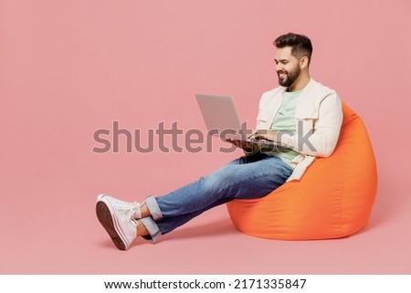 Full body young smiling happy man in trendy jacket shirt sit in bag chair hold use work on laptop pc computer isolated on plain pastel light pink background studio portrait. People lifestyle concept