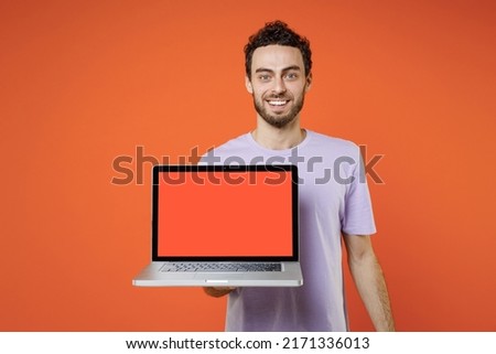 Smiling young bearded man 20s wearing casual basic violet t-shirt standing hold laptop pc computer with blank empty screen looking camera isolated on orange color wall background studio portrait