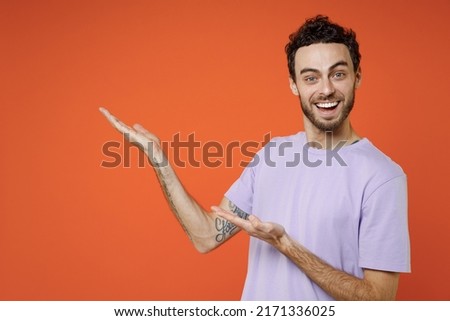 Smiling cheerful young bearded man 20s wears casual violet t-shirt standing pointing hands aside on mock up copy space looking camera isolated on orange background studio portrait Tattoo translate fun