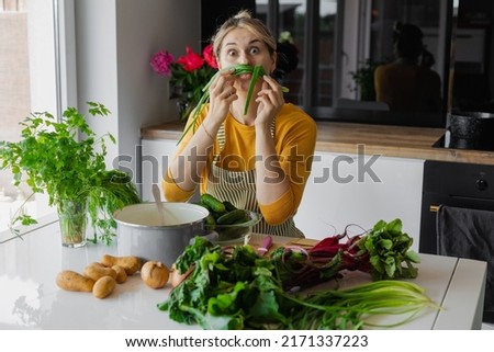 Playful and funny blonde woman holding sprout onion like mustache and looking at camera. Humorous cooking of mixed eco vegetable salad at home. Healthy eating and organic food diet in summer. 