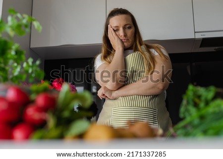 Tired, sad and bored plus size blond woman holding head in hand, thinking what to cook, have no idea. Diet and healthy eating with vegetables. Home cooking, looking for recipe. Greenery on foreground