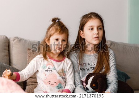 Two little girls sisters wear pyjamas sitting on beige couch at home with plush soft toys,  resting, having fun together. Familytime at weekend