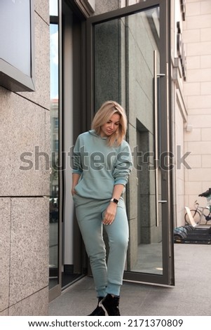 teen walk leisure. young blonde girl in mint oversize costume with hoodie is standing casual near street store door background and looking down with cute smile. lifestyle concept, free space