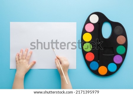 Baby hand holding paint brush. Colorful palette and empty white paper on light blue table background. Pastel color. Closeup. Point of view shot.