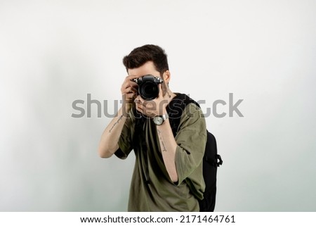 Portrait of young caucasian man wearing t-shirt posing isolated over white background taking images with dslr camera. Photographer covering his face with the camera.