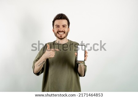 Cheerful caucasian man wearing casual clothes posing isolated over white background showing to camera and pointing at device. Gadget with empty free space for mock up. White blank screen.
