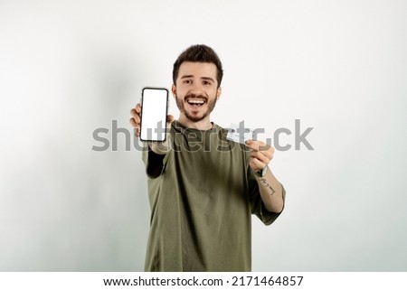 Cheerful caucasian man wearing casual clothes posing isolated over white background holding credit card in hand and showing phone with white blank screen to camera. Shopping or online payment, banking