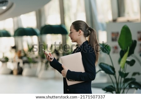 A photo from the side of female realtor who is holding a laptop and scrolling a smartphone. A woman is standing in the modern lobby. A businesswoman is leaving her office in a contemporary interior.