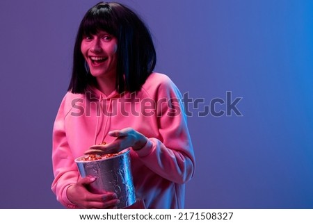 Laughing awesome brunet woman in pink hoodie with popcorn look at camera hold palm up posing isolated in blue violet color light studio background. Neon party Cyberpunk Cinema concept. Copy space