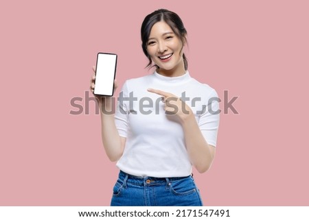 Attractive Young Asian woman pointing finger to mobile smartphone blank screen on isolated pink background.