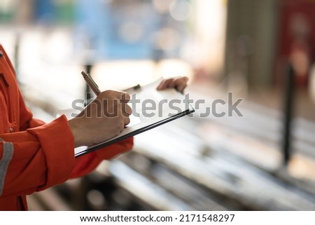 Action of safety officer is writing on safety checklist document during safety audit and inspection, with factory workshop as blurred background. Industrial expertise occupation photo. Selective focus Royalty-Free Stock Photo #2171548297