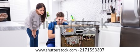Smiling Woman Behind Technician Repairing Dishwasher In Kitchen