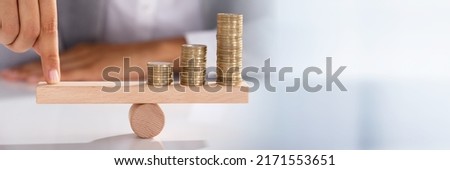 Close-up Of Businessperson's Finger Balancing Stacked Of Coins On Wooden Seesaw