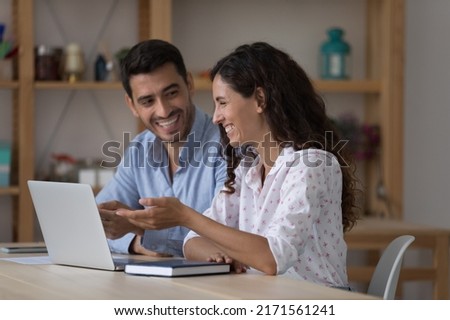 Attractive laughing Hispanic colleagues sit at desk with laptop work on collaborative task, on-line project looking cheerful, joking while take break. Teamwork, friendship at workplace, tech concept Royalty-Free Stock Photo #2171561241