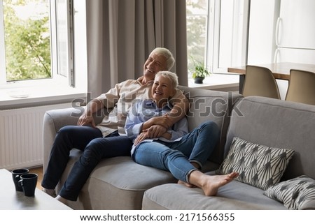 Joyful older grey-haired couple in love hugging relaxing on sofa in living room laughing while watching comedy movie on TV. Happy carefree spouses talking feel overjoyed spend weekend leisure at home Royalty-Free Stock Photo #2171566355