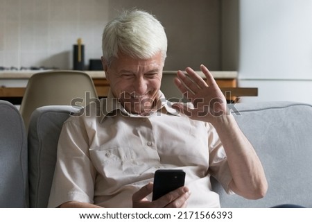 Overjoyed senior man sits on sofa at home staring at smart phone screen greets grown up children living abroad using video conference app on modern cellphone. Virtual meeting, comfort, tech concept Royalty-Free Stock Photo #2171566393