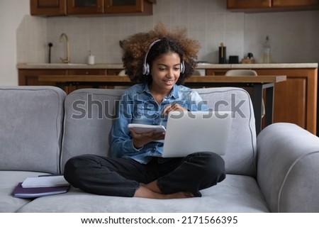Cute teenage girl wear headphones makes assignment sit on sofa with laptop, gaining new knowledge use internet, listen audio course, improve foreign language skills. Video call, study at home concept