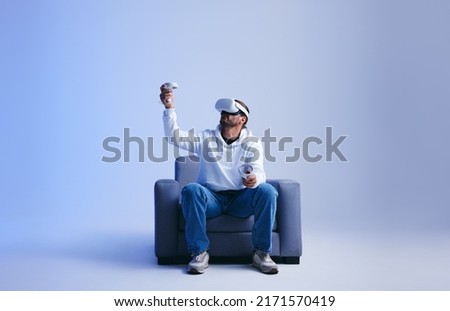 Young man playing an immersive virtual reality game on a couch. Man gaming with virtual reality goggles and controllers. Young man experiencing a 3D simulation. Royalty-Free Stock Photo #2171570419
