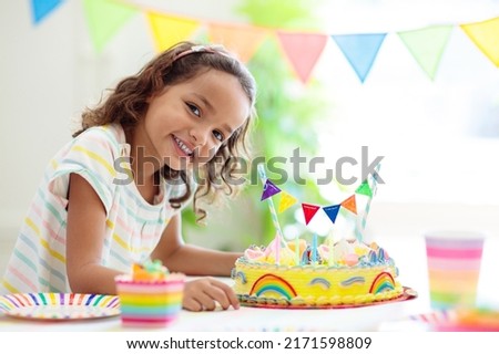 Kids birthday party. Children celebrate with colorful cake and gifts. Little curly girl blowing candles and opening birthday presents. Friends play with rainbow confetti. Party home decoration.