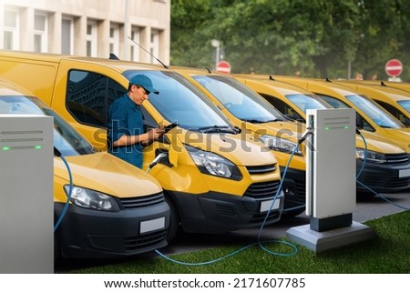 A man with a digital tablet stands next to yellow electric delivery vans at electric vehicle charging stations Royalty-Free Stock Photo #2171607785