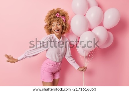 Positive surpised woman spends free time on welcome party holds bunch of inflated balloons wears blouse and shorts feels very happy dances against pink background. Birthday celebration concept Royalty-Free Stock Photo #2171616853