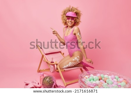 Summer vibes. Happy woman with curly hair applies sunscream to protect skin from sunburn poses on lounge chair points into distance isolated over pink background. Leisure and recreation concept