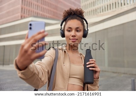 Horizontal shot of sporty active woman with curly combed hair takes selfie via smartphone holds bottle with fresh water rests after running or cardio training dressed in activewear poses outdoors