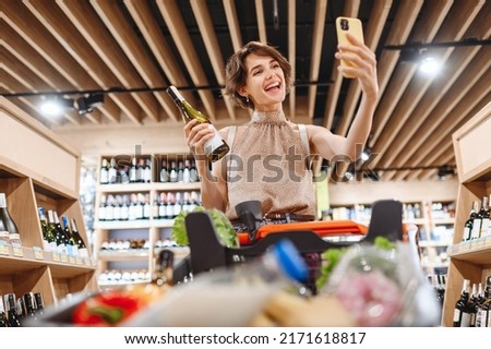 Young fun woman in casual clothes shopping at supermaket store with grocery cart hold white wine alcohol hold bottle do selfie shot on mobile phone inside hypermarket Purchasing gastronomy concept.
