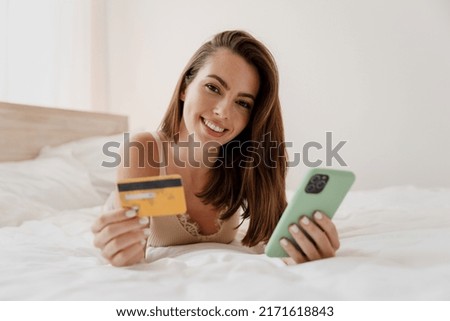 Satisfied fun young woman in casual casual clothes lying on her stomach hold credit bank card doing online shopping look camera rest relax spend time in bedroom lounge home in own room house good day