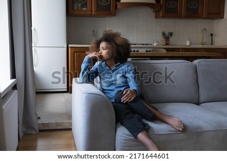 Sad African teen girl sit on sofa lost in thoughts, thinks staring into distance, looks upset experiences first unrequited love, having low self-esteem, problems, feels insecure. Teen problem concept Royalty-Free Stock Photo #2171644601