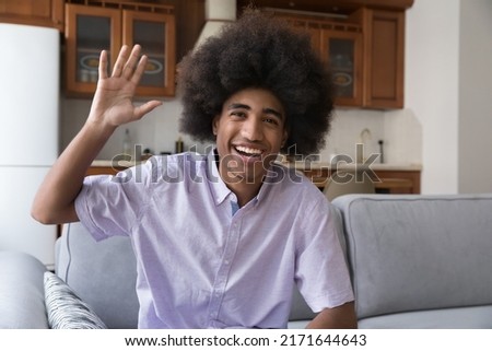 Happy teenage African curly-haired guy sit on couch smile look at camera, wave hand greets friend start videocall. Electronic dating, distance communication, young gen and modern tech usage concept Royalty-Free Stock Photo #2171644643
